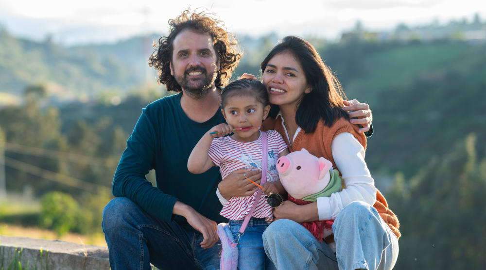 A family of three sits on a small stone fence and smiles at the camera.